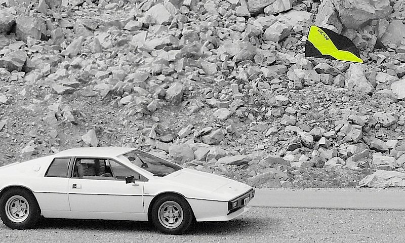 A neon yellow light wind kite is floating above a white Lotus Esprit.